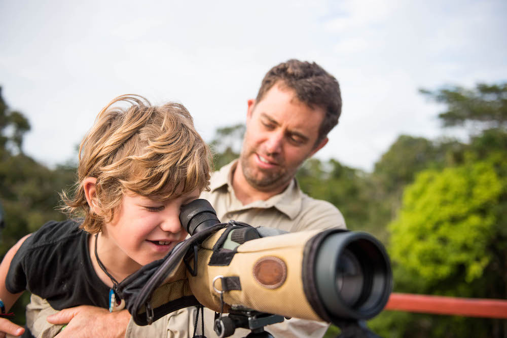 little boy being held by adult as he looks into a telescope at a distant animal